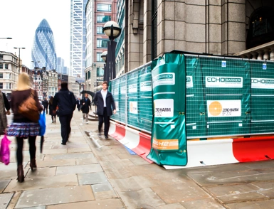 Green construction hoarding and Echo safety barriers on a busy London street near the Gherkin