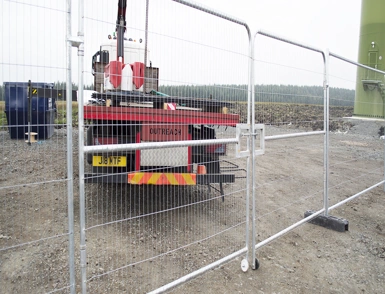 Temporary metal security fencing and gate at a construction site, with a crane truck behind it near a large green turbine tower base