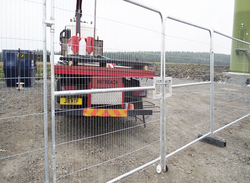 Temporary metal security fencing and gate at a construction site, with a crane truck behind it near a large green turbine tower base